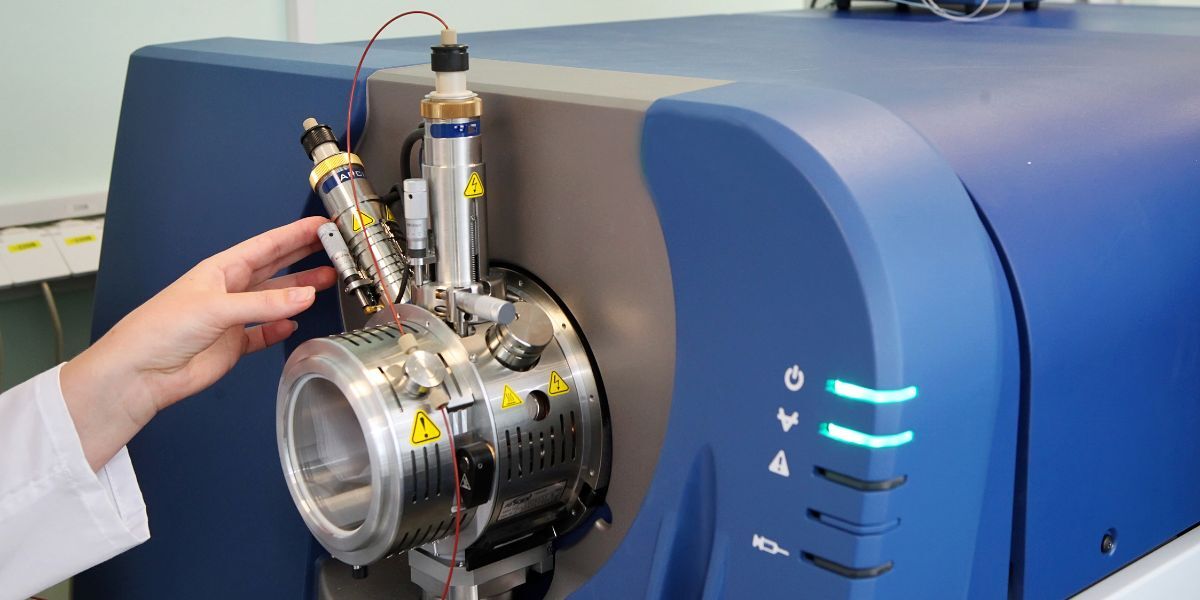 A technician’s hand adjusts components on a large blue mass spectrometer, highlighting the complex hardware involved in repair and maintenance decisions.