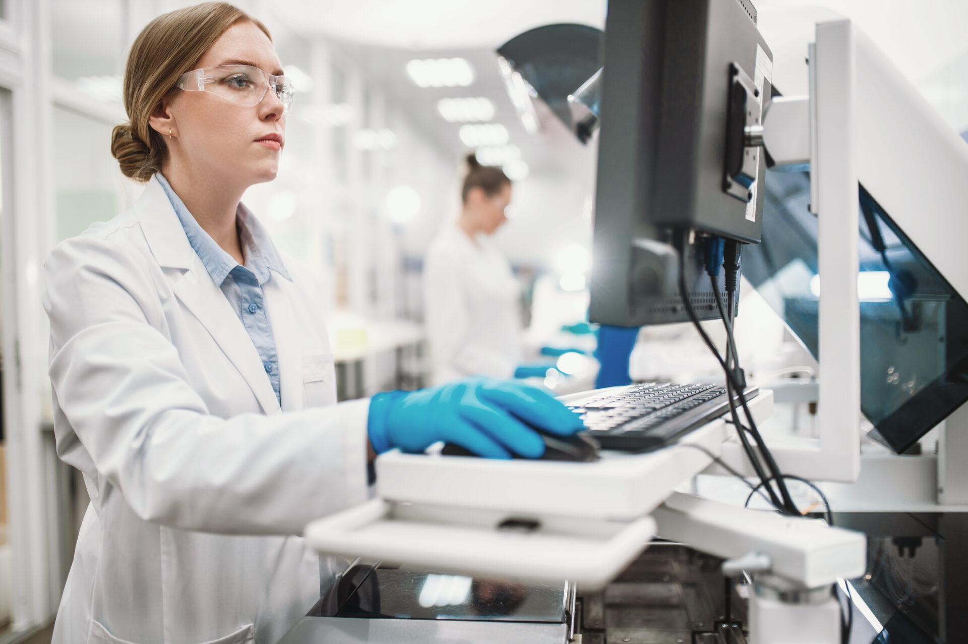 A female laboratory technician wearing a white lab coat, safety glasses, and blue nitrile gloves types on a keyboard while working at a computer workstation in a bright, modern laboratory. A second lab worker is visible in the blurred background.